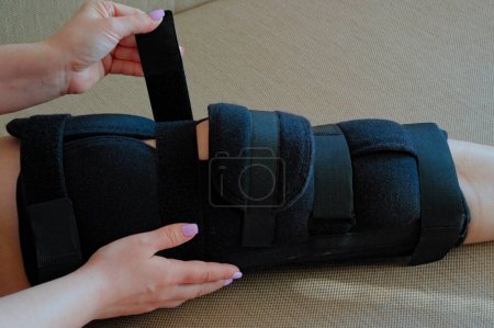 Foto de Mujer joven que fija la ortesis ajustable en la pierna rota sentada en la cama. Usar corsé de pierna después de lesionarse. Vista de cerca. Rehabilitación de la mujer lesionada en casa. - Imagen libre de derechos