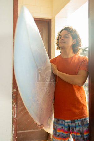 Vertical photo of a young casual latin repairman carrying a surfboard into a workshop