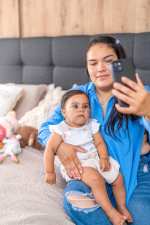A relaxed mother holding her baby takes a selfie with a smartphone, capturing everyday family moments.