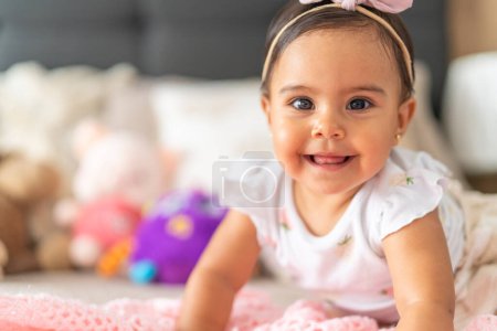 A baby girl with a radiant smile lies on a pink blanket, surrounded by plush toys, in a bright and cheerful setting.