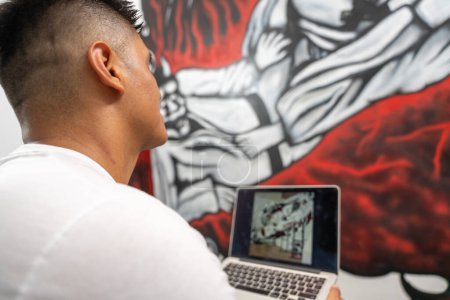 Rear view of a young latin male muralist using laptop while working in a new mural indoors
