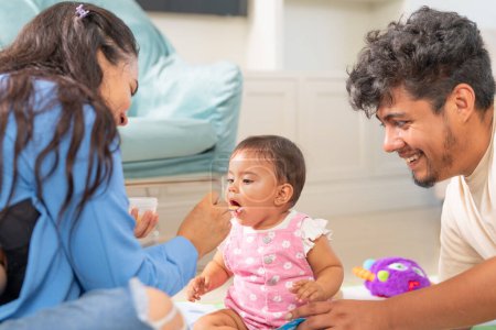 A joyful family moment as a baby girl is fed by one parent, with the other parent smiling encouragingly during mealtime.