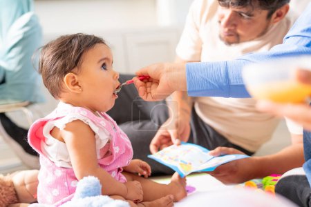 A baby girl eagerly opens her mouth for a spoonful of food during a family mealtime, with a parent engaged in storytelling.