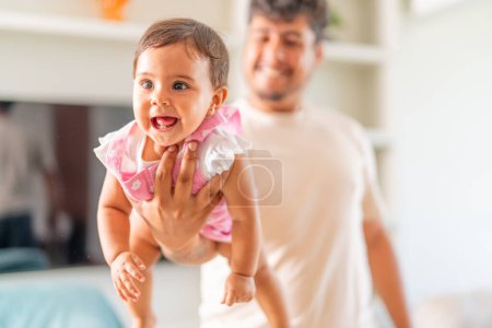 A father lifts his baby daughter in the air, both enjoying a moment of laughter and play in a bright, home setting.