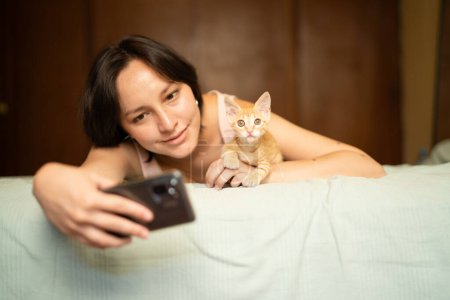 A cozy scene of a young woman lying down taking a selfie with her curious ginger kitten, sharing a moment of connection.