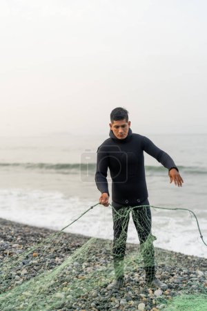 Retrato vertical de un joven pescador latino preparando una red en la playa en un día nublado
