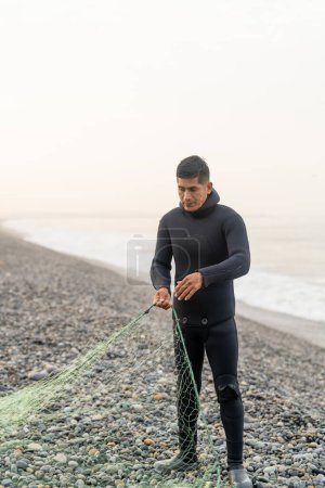 Foto vertical de un pescador latino con traje de neopreno preparando una red en la playa en un día nublado