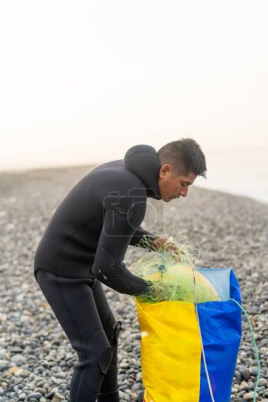 Fotografía vertical con espacio de copia de un joven pescador latino con traje de neopreno sacando una red de una bolsa de plástico en la playa