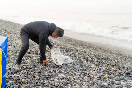 Hombre con traje de neopreno preparando una bolsa con piedras para flotar mientras pesca en la playa en un día nublado