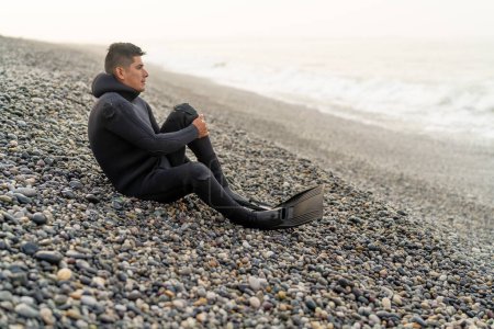 Joven pescador latino sentado en la playa con traje de neopreno y aletas en un día frío