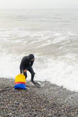 Vertical photo of a fisherman with wetsuit and fins carrying a heavy bag with stones and net to fish
