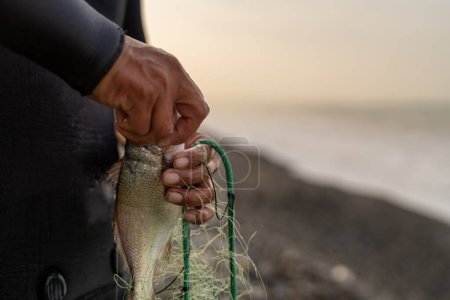 Primer plano de las manos de un pescador irreconocible en traje de neopreno recogiendo peces de una red de pie en la playa