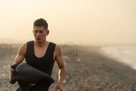 Foto con espacio de copia de un joven pescador latino fuerte quitándose el traje de neopreno en la playa