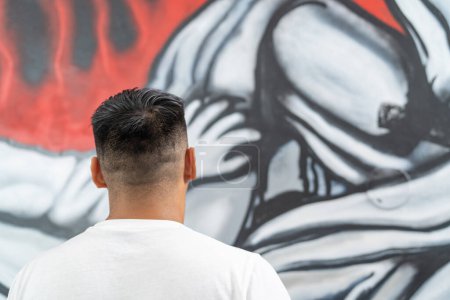 Vista trasera de un joven artista latino mirando su pintura en la pared