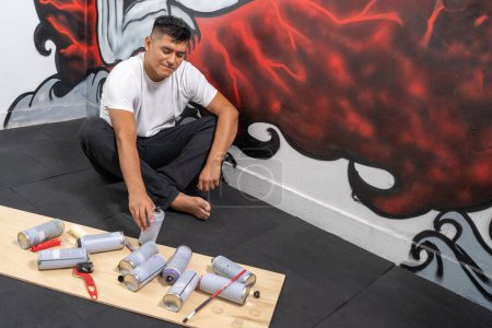 Téléchargez les photos : Vue surélevée d'un jeune peintre de rue latino assis à choisir des aérosols à côté d'une nouvelle murale - en image libre de droit