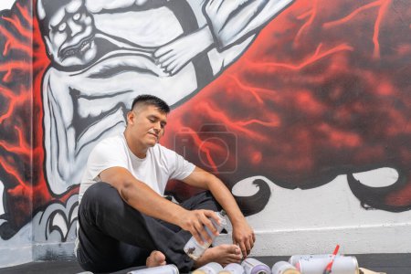 Horizontal photo with copy space of a latin young male tired muralist taking a break from work sitting on the floor