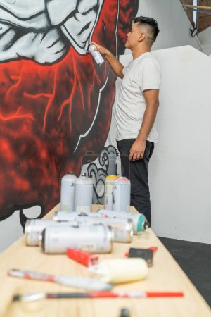 Foto vertical de un muralista latino joven trabajando usando spray en un nuevo mural en el interior