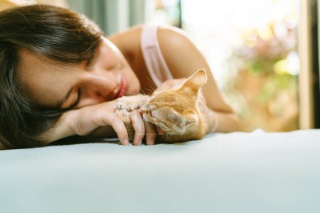 A woman lies close to a ginger kitten, gently holding its paw, in a display of affection and companionship.