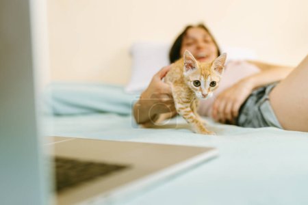 A woman relaxes on a bed, smiling as she interacts with a playful ginger kitten, with a laptop in the foreground suggesting a casual home office setting.