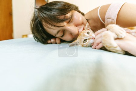 Une femme s'allonge avec son visage près d'un chaton roux, partageant un moment tendre et aimant sur un couvre-lit bleu doux.