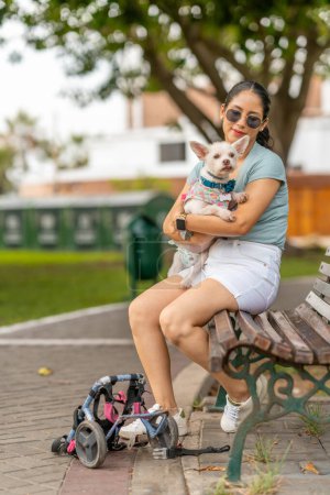 Eine Frau hält ihren behinderten Hund liebevoll in der Hand und macht eine Pause von der Benutzung ihres Rollstuhls im Park.