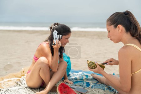 Mujer latina joven escucha como la película de la cámara se ha agotado en la playa mientras toma fotos durante un picnic con un amigo