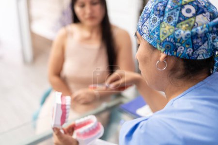 A dentist in a patterned cap demonstrates toothbrushing on a dental model for a focused patient in a clinic