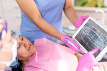 In a bright dental office, a dentist shows a panoramic dental X-ray to a patient on a tablet while she reclines in the dental chair