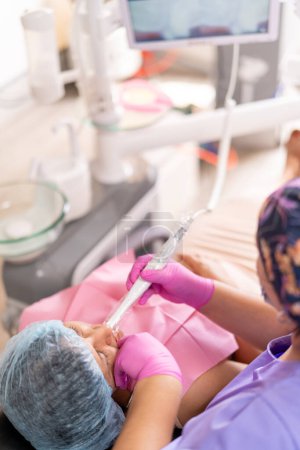 Un dentista con un uniforme púrpura y guantes rosados realiza un procedimiento dental en un paciente reclinado, con equipo dental en el fondo