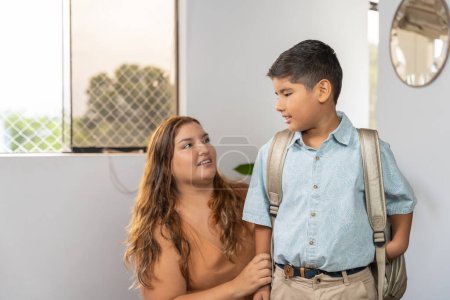 Madre latina con sobrepeso e hijo pequeño con uniforme preparándose para el primer día en la escuela en casa