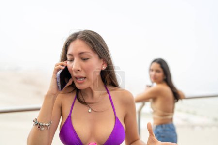 Mujer discute mientras habla por teléfono mientras un amigo observa de pie en un balcón con vistas al mar