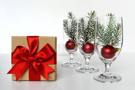 Photo for Christmas decorations on the table, gift box with red ribbon and champagne glasses with red shiny balls and spruce twigs - Royalty Free Image