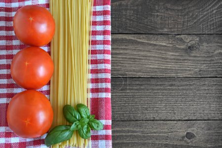 Foto de Antecedentes de comida italiana. tomates frescos, hojas de albahaca, espaguetis crudos y paño a cuadros en mesa de madera con espacio para copiar - Imagen libre de derechos