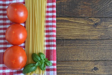 Foto de Antecedentes de comida italiana. tomates frescos, hojas de albahaca, espaguetis crudos y tela a cuadros sobre fondo de madera. Copiar espacio - Imagen libre de derechos