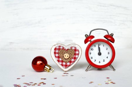 Foto de Composición navideña. bauble rojo, decoración del corazón de madera y reloj despertador vintage con confeti sobre fondo de madera blanco - Imagen libre de derechos