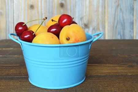 Photo for Sweet fresh apricots and cherries in blue bucket on wooden table - Royalty Free Image