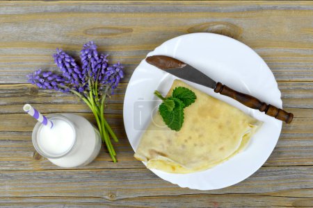 Foto de Panqueque y cuchillo en plato blanco, botella de leche y flores violetas en mesa de madera, vista superior - Imagen libre de derechos