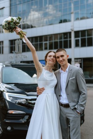 Téléchargez les photos : Mariée et marié avec une voiture noire près d'un gratte-ciel en verre - en image libre de droit