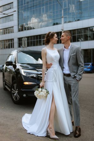 Brautpaar mit schwarzem Auto in der Nähe eines gläsernen Wolkenkratzers