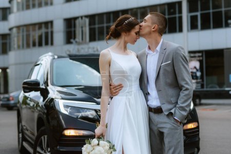 Téléchargez les photos : Mariée et marié avec une voiture noire près d'un gratte-ciel en verre - en image libre de droit