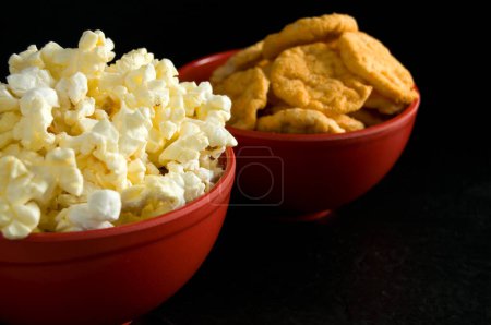 Photo for Close Side View of Buttered Popcorn and BBQ Rice Chips in Red Bowls Isolated on Black Horizontal - Royalty Free Image