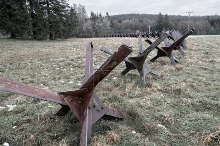 Foto de Old steel rusty anti tank roadblocks laid on grass with forest in the background on a gloomy dark day of winter without snow. Concept for war, defense, hard times - Imagen libre de derechos