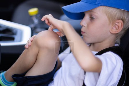 Kleiner Junge, der im Auto sitzt, auf seine Knie schaut und trockene Haut von der Wunde befreit. Schuljunge mit Schorf auf den Knien