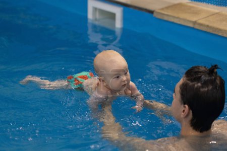 Baby schaut interessiert zum Schwimmtrainer. Schwimmtrainer hält Neugeborenes in der Hand, bringt ihm Schwimmen bei. 