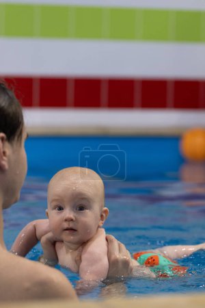 Porträt des Babys in einem lustigen Badehöschen mit einem Schwimmtrainer in einem Schwimmbad. Schwimmunterricht für Neugeborene. Nahaufnahme