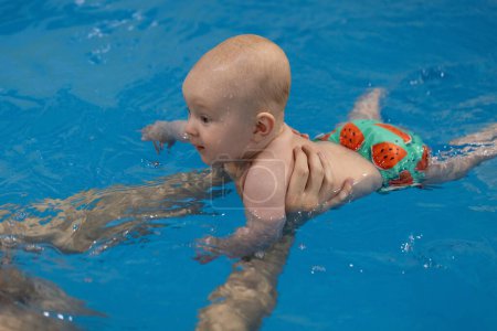 Überraschtes Baby schwimmt auf blauem Wasser im Schwimmbad. Mans Hände helfen dem Baby, auf dem Wasser zu schwimmen. Nahaufnahme.