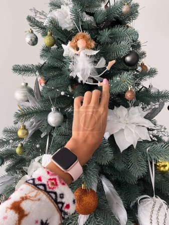 Téléchargez les photos : Main féminine avec montre intelligente touchant petit ange blanc sur l'arbre de Noël. Gros plan. - en image libre de droit