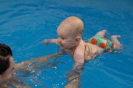 Lächelndes Baby im lustigen Badehöschen, das mit Vater im Schwimmbad schwimmt. Schwimmen mit Neugeborenen. 