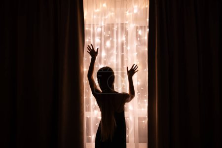 Téléchargez les photos : Silhouette sombre d'une femme debout près de la fenêtre avec éclairage de Noël - en image libre de droit
