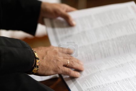 Hands of the priest on the pages of the holy book. Priest in the black robe reading names of the saints before christianity, close up